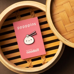 Puede incluir: Una tarjeta rosa con el texto "pppppins" y un pin de dumpling con palillos chinos descansa dentro de una vaporera de bambú. El pin es blanco con detalles negros y una expresión linda. La tarjeta también dice "Designed by noodoll".