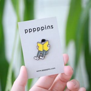 May include: A gray card with the text "pppppins" and a pin of a person reading a yellow book. The pin is black, white, yellow, and gray. The website www.pppppins.com is printed at the bottom of the card.