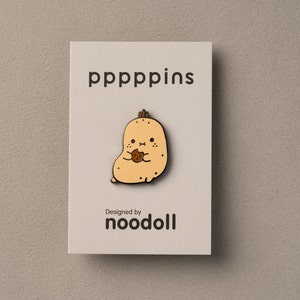 May include: A tan enamel pin featuring a cartoon potato character holding a cookie. The pin is on a light beige card with the text "pppppins" and "Designed by noodoll" in black.