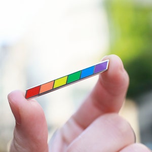Puede incluir: Un pin rectangular esmaltado con un esquema de color arcoíris, sostenido entre los dedos. El pin presenta un borde plateado y una serie de rectángulos de colores: rojo, naranja, amarillo, verde, azul y morado. El fondo está borroso.