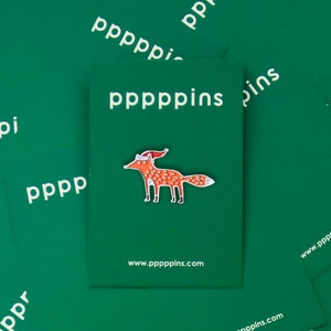 May include: A colorful fox enamel pin with a Santa hat, set against a green background with the text "pppppins" and the website address. The pin is orange, white, and red, and is on a green card.