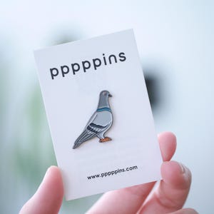 Peut inclure: Épingle en émail en forme de pigeon, avec des détails gris, bleus et orange, présentée sur une carte blanche. La carte porte le texte "pppppins" et l'adresse du site web www.pppppins.com.