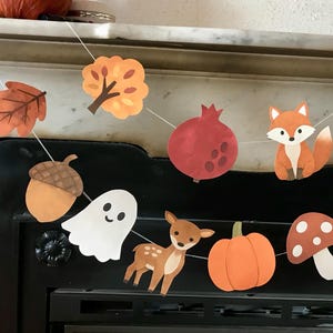 May include: A decorative garland featuring paper cutouts of autumn-themed icons including a fox, a deer, a ghost, a pumpkin, an acorn, a pomegranate, a hedgehog, mushrooms, an apple, and fall leaves, all strung together on a thin white cord.