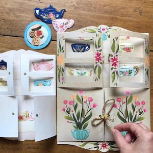 May include: A paper craft set featuring two folding cabinet structures with open shelves displaying illustrations of teapots, teacups, and plates. The larger cabinet features floral designs and a gold glittery string closure, while loose paper cutouts of a teapot, teacup, and cookies are included.
