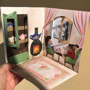May include: A pop-up greeting card featuring an illustrated interior scene with a green cabinet, a fireplace with a blue teapot, and two anthropomorphic animals sitting in green chairs by a window showing a snowy landscape and a pink rug.