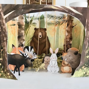 Peut inclure: Un diorama en papier multicouche représentant une scène de forêt avec un ours brun, un élan, un lapin, un hérisson, un blaireau, une tortue et un sanglier. La scène est encadrée par des arbres et du feuillage, créant un effet tridimensionnel.