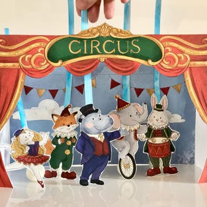 Op de afbeelding: Een papieren circuspodium met het woord CIRCUS in gouden letters op een groene banner. De scène bevat vijf geïllustreerde dierenfiguren: een kat in een rood tutu, een vos, een nijlpaard in een pak, een olifant op een eenwieler en een konijn.
