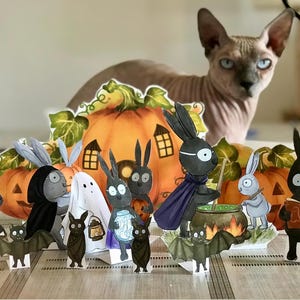 May include: A collection of paper cutouts featuring black rabbits dressed in costumes, a ghost, and bats arranged in front of a large orange pumpkin backdrop. The scene includes a rabbit stirring a green cauldron and another reading a book.