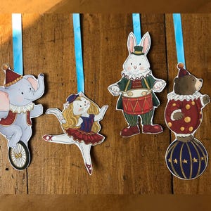 Può includere: Quattro ornamenti pendenti con illustrazioni a tema circense: un elefante grigio su un monociclo, una gatta ballerina bionda, un coniglio bianco che suona un tamburo rosso e un orso bruno in equilibrio su una palla blu, con nastro azzurro.