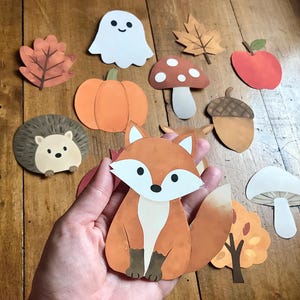 Könnte beinhalten: Eine Sammlung von Papier-Ausschnitten mit herbstlichen Illustrationen, darunter ein Fuchs, ein Geist, ein Kürbis, ein Igel, ein Pilz, eine Eichel, ein Apfel und verschiedene Blätter. Der Fuchsausschnitt ist orange, weiß und braun gefärbt.