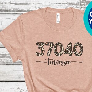 May include: A peach-colored t-shirt with the text "37040 Tennessee" in black script and leopard print. A blue circle with the text "CUSTOMIZE YOUR SHIRT" is in the upper right corner. The shirt is knotted at the bottom.