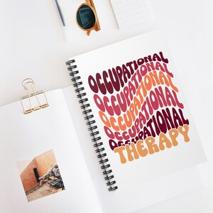 May include: A white spiral-bound notebook with the words "OCCUPATIONAL THERAPY" in a retro, wavy font. The text is in shades of burgundy, orange, and peach. The notebook is open, and a gold binder clip and sunglasses are visible.