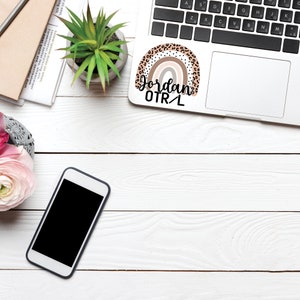 May include: A white laptop with a rainbow sticker that says "Jordan OTR/L" on the top right corner. The laptop is on a white wooden surface with a potted plant, a cell phone, and a notebook.