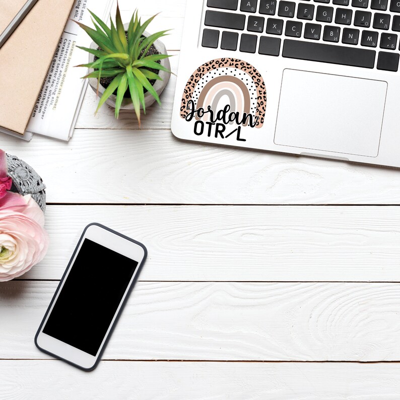May include: A white laptop with a rainbow sticker that says "Jordan OTR/L" on the top right corner. The laptop is on a white wooden surface with a potted plant, a cell phone, and a notebook.
