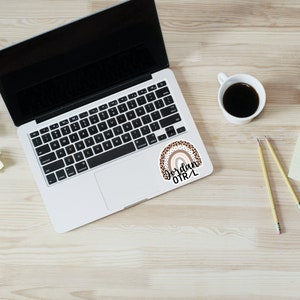 May include: A sticker with a rainbow design in shades of brown and pink with black leopard print accents. The rainbow has a white outline and the text "Jordan OTRL" is written in black cursive below the rainbow.