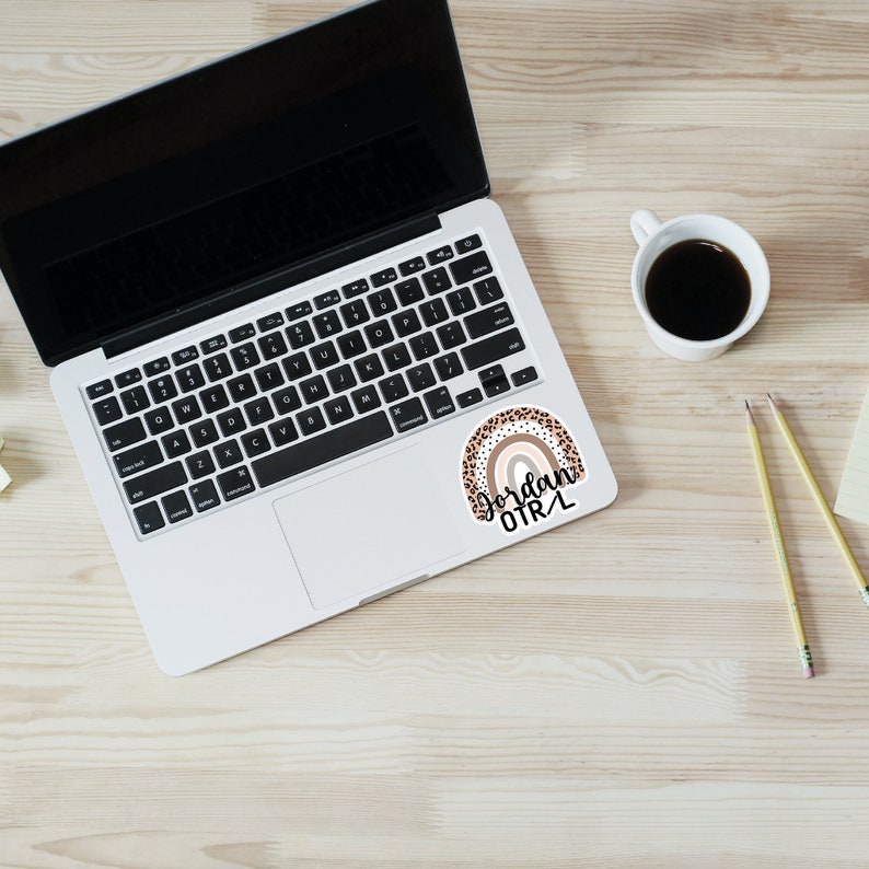 May include: A sticker with a rainbow design in shades of brown and pink with black leopard print accents. The rainbow has a white outline and the text "Jordan OTRL" is written in black cursive below the rainbow.
