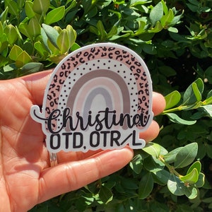 May include: A white sticker with a rainbow design and black leopard print border. The sticker says "Christina OTD, OTR/L" in black text.