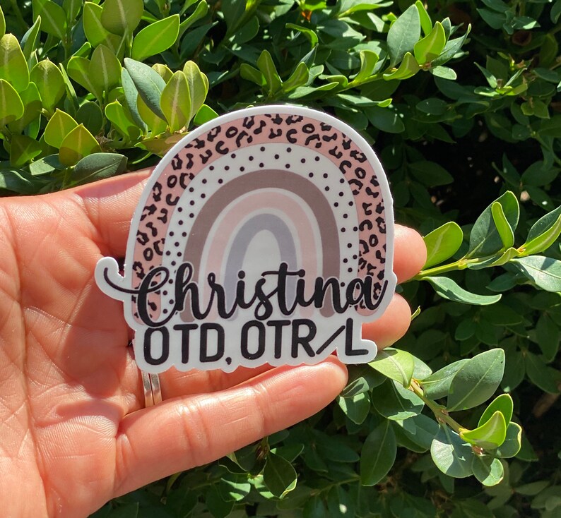 May include: A white sticker with a rainbow design and black leopard print border. The sticker says "Christina OTD, OTR/L" in black text.