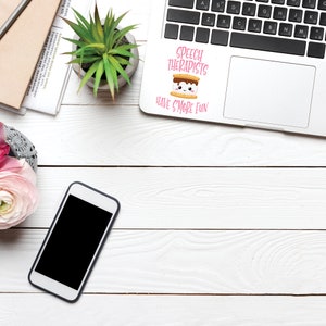 May include: A white laptop computer with a pink and brown sticker that says "Speech Therapists Have S'more Fun". The sticker features a cartoon marshmallow with a smiling face. The laptop is on a white wooden surface with a potted plant, a cell phone, and a notebook.