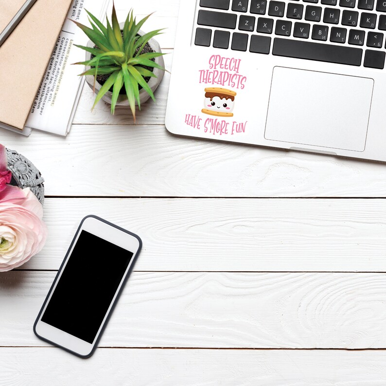 May include: A white laptop computer with a pink and brown sticker that says "Speech Therapists Have S'more Fun". The sticker features a cartoon marshmallow with a smiling face. The laptop is on a white wooden surface with a potted plant, a cell phone, and a notebook.