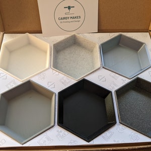 May include: Six hexagonal-shaped containers made of different colours of plastic. The containers are arranged in a honeycomb pattern. The containers are in a box with a white background and the text "CAIRDY MAKES 3D Printing and Design" printed on the top.