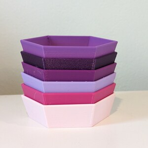 May include: A stack of six hexagonal-shaped containers in shades of purple, pink, and lavender. The containers are made of plastic and are ideal for storing small items.