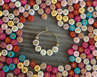 May include: A gold hoop earring with the word "BRIDE" spelled out in white letter beads. The earring is surrounded by a collection of colorful round beads with gold letters.