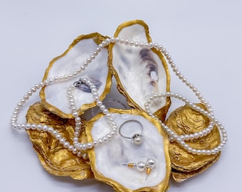Puede incluir: Un conjunto de cuatro conchas de ostra pintadas de oro con un collar de perlas, un anillo y un par de pendientes. Las conchas están dispuestas en forma de pirámide.