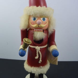 Peut inclure: Un casse-noisette en bois représentant le Père Noël avec un chapeau rouge, une barbe blanche et une cloche dorée dans la main. Il porte une robe rouge avec une bordure en fourrure blanche et une ceinture blanche. Il est debout sur une base blanche.