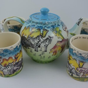 May include: A set of four ceramic mugs and a teapot with a blue lid. The mugs and teapot are decorated with a whimsical animal print featuring zebras, hippos, and other animals. The text on the mugs reads "We went in two by two".