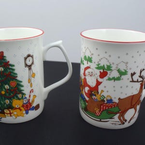 May include: Two white ceramic mugs with red rim accents. The left mug features a decorated Christmas tree, a wall clock, and toys. The right mug displays Santa Claus in a green sleigh pulled by a reindeer, surrounded by snow-covered trees.