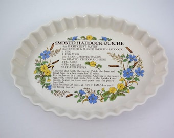 May include: A white ceramic baking dish with a scalloped edge and a floral design. The dish has a recipe for Smoked Haddock Quiche printed on it, including ingredients and instructions.