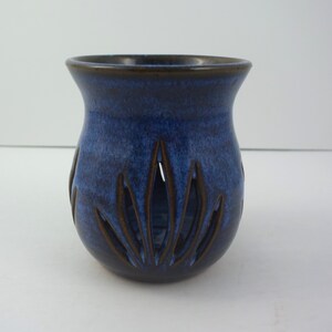 May include: A ceramic candle holder with a deep blue and black mottled glaze. The vessel features a wide, flared rim and a rounded body with vertical, petal-shaped cutouts around the base that reveal the dark brown interior clay surface.