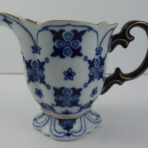 May include: A white and blue porcelain creamer with a floral pattern. The creamer has a scalloped rim and base, a dark blue handle with gold accents, and a spout. The creamer is approximately 13 cm tall.