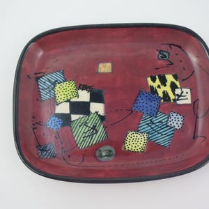May include: A rectangular ceramic tray with a deep red base and a black rim. The surface features scattered square patterns in yellow, blue, and black, including a checkerboard design and various dotted or striped textures, with the text KBCS visible.