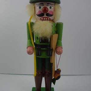 May include: A wooden nutcracker figurine of a hunter wearing a green hat, green jacket, and brown trousers. He is holding a rifle and binoculars. The nutcracker is standing on a green base.