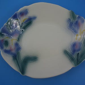 May include: White oval ceramic dish with a blue and green floral design. The dish features two stylised iris flowers with yellow centres and green leaves.