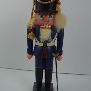 Peut inclure: Une figurine de casse-noisette en bois habillée en soldat. Il porte un uniforme bleu avec des accents rouges, une ceinture blanche et un chapeau noir avec des garnitures dorées. Il a une barbe et une moustache blanches et tient un bâton noir et doré.