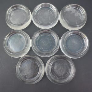 May include: A collection of eight circular, clear glass coasters arranged in a grid pattern. Each coaster features a smooth, rounded rim and a flat base with the embossed text "MADE IN U.S.A." visible on the bottom surface of several pieces.