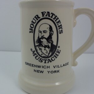 Peut inclure: Une tasse en céramique blanche avec une illustration en noir et blanc d'un homme avec une moustache. Le texte "Your Father's Mustache" est dans un cercle autour de l'illustration. Le texte "Greenwich Village New York" est sous le cercle.
