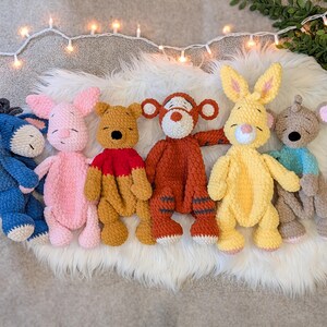 May include: A collection of six handmade crochet plush toys featuring a blue donkey, a pink pig, a yellow and red bear, an orange and black striped tiger, a yellow rabbit, and a tan mouse with a blue shirt, all arranged in a row on a white surface.