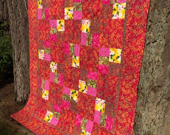 May include: A red quilt with a patchwork design featuring squares of pink, yellow, and green floral fabric. The quilt is hanging on a tree trunk.
