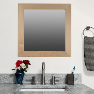 May include: A square, light-wood framed mirror hangs above a bathroom sink with a brushed nickel faucet. A dark blue vase with red and white flowers sits on the countertop. A gray towel hangs from a metal ring.