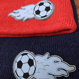 May include: Two knit beanies with a soccer ball and flame design. The top beanie is red and the bottom beanie is navy blue.