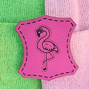 May include: A pink leather patch with a black flamingo design, stitched onto a green and pink knitted fabric. The flamingo is standing on one leg. The patch has a decorative stitched border.
