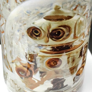 May include: White ceramic mug with a colorful illustration of a robot with large eyes and a gold-colored body. The mug has the word "THANK" printed on the bottom.