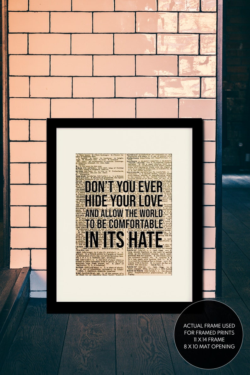 Puede incluir: Una impresi&oacute;n enmarcada en negro con un fondo de peri&oacute;dico vintage que presenta el texto "Don't you ever hide your love and allow the world to be comfortable in its hate". La impresi&oacute;n est&aacute; enmarcada con un marco y un passe-partout negros, y el tama&ntilde;o real del marco es de 28 x 35,5 cm con una abertura de passe-partout de 20,3 x 25,4 cm.