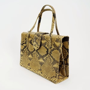Può includere: Borsa a mano rettangolare con motivo a pelle di serpente nei toni del beige, marrone e nero. La borsa è dotata di chiusura con patta e due manici superiori. La borsa è realizzata in un materiale strutturato.