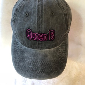 May include: A gray, washed-out baseball cap with the embroidered text "Queen B" in pink. The cap has a curved brim and a button on top. The hat is made of a soft, textured material.