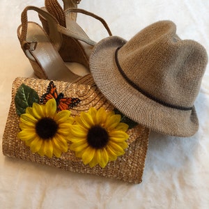 May include: A collection of summer accessories including a woven straw clutch with sunflower and butterfly embellishments, a pair of wedge sandals, and a tan straw hat. The clutch features two large yellow sunflowers and a monarch butterfly.